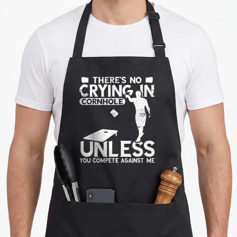 Crying game aprons - Distressed white silhouette of person throwing bean bag with cornhole board and sarcastic text — shown on a black cotton canvas apron against a white background