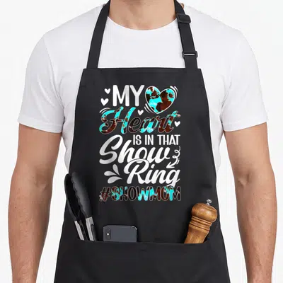 Ring cooking mama apron - Stacked text My Heart IS IN THAT Show Ring with cow print heart and hashtag — shown on a black cotton canvas apron against a white background