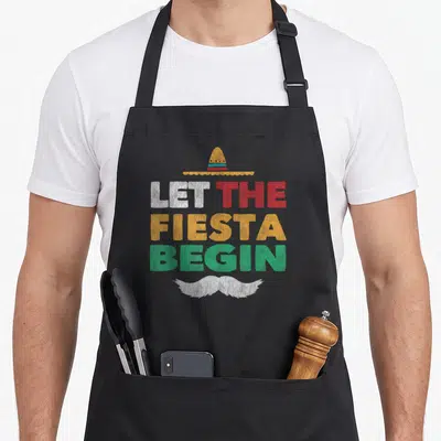 Fiesta cinco mayo mexican apron - Distressed text LET THE FIESTA BEGIN with sombrero above and mustache below in white — shown on a black cotton canvas apron against a white background