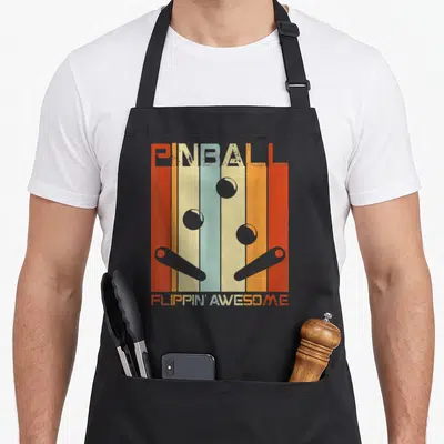 Machine game aprons - Retro pinball art with vertical stripes, three balls, two flippers, and text PINBALL FLIPPIN' — shown on a black cotton canvas apron against a white background