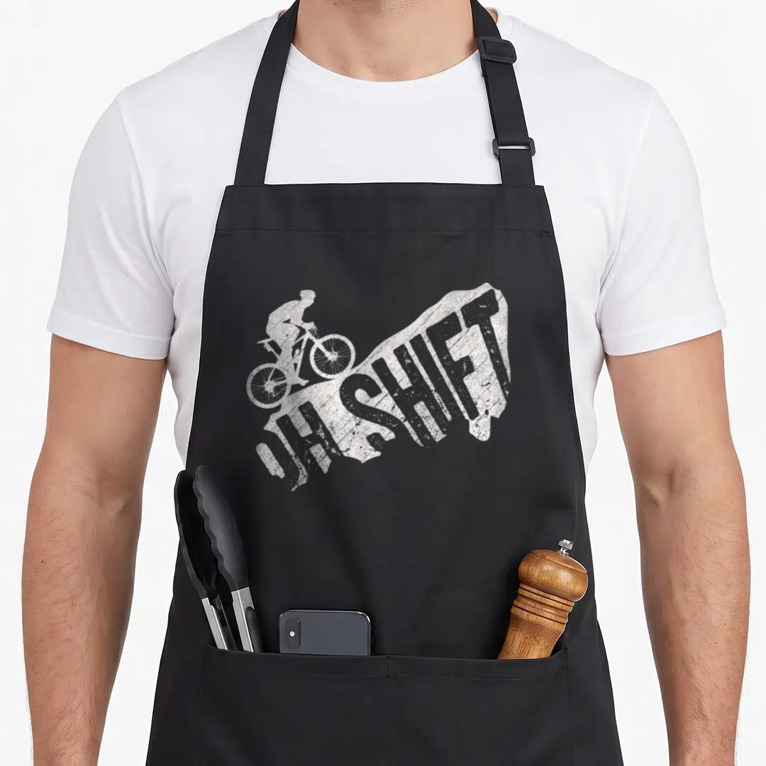 Shift bicycle apron - Distressed white mountain biker silhouette riding over OH SHIFT text on a dirt mound shape — shown on a black cotton canvas apron against a white background
