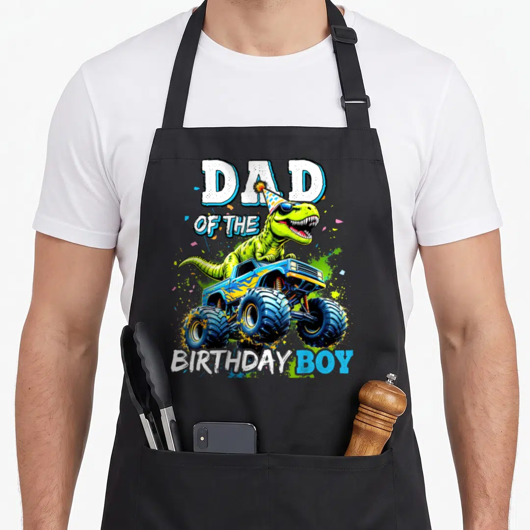Dinosaur rex truck apron - Green T-Rex in party hat driving blue monster truck with Dad of the Birthday Boy text — shown on a black cotton canvas apron against a white background