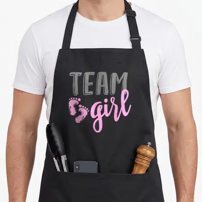 Baby gender reveal apron - Pink baby footprints and girl text on grey background with faint TEAM letters — shown on a black cotton canvas apron against a white background
