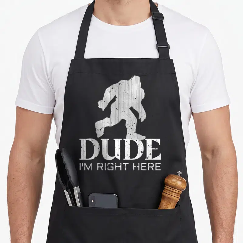 Dude yeti apron - White distressed bigfoot silhouette above stacked text DUDE I'M RIGHT HERE — shown on a black cotton canvas apron against a white background