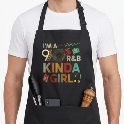 Kinda throwback r aprons - Stacked text I'M A 90'S R&B KINDA GIRL with vinyl, TV, cassette, headphones in beige orange — shown on a black cotton canvas apron against a white background