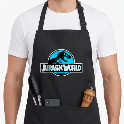 Blue science apron - JURASSIC WORLD text over black T-Rex skeleton in blue circle with palm trees — shown on a black cotton canvas apron against a white background