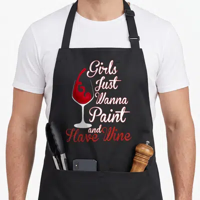 Wine sip and paint aprons - White wine glass with red splash and stacked text reading Girls Just Wanna Paint and Have — shown on a black cotton canvas apron against a white background