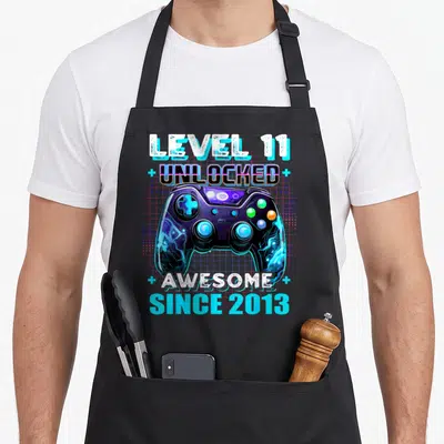 Eleven birthday apron - Retro game controller with Level 11 Unlocked text and pink grid background — shown on a black cotton canvas apron against a white background