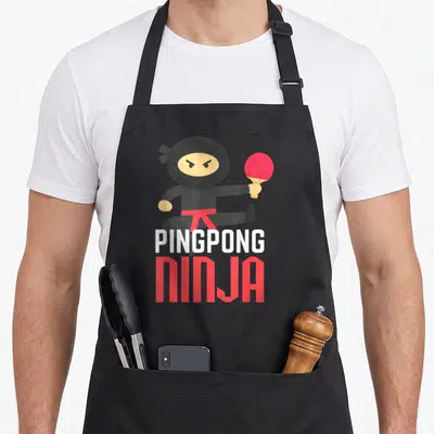 Ping table apron - Cartoon ninja holding paddle and ball above PINGPONG NINJA text in black, red, white, yellow — shown on a black cotton canvas apron against a white background