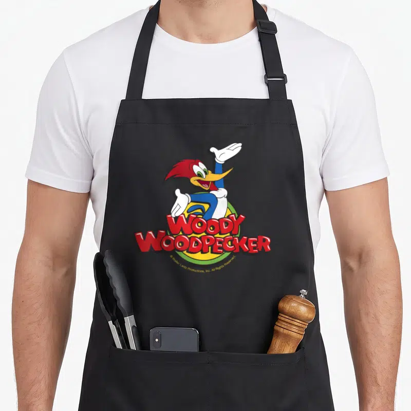 Funny woodpecker apron - Woody Woodpecker cartoon character in red and blue over yellow circle with red text — shown on a black cotton canvas apron against a white background
