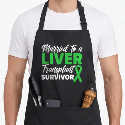 Married liver organ apron - White and green text Married to a LIVER Transplant SURVIVOR with green ribbon on right side — shown on a black cotton canvas apron against a white background