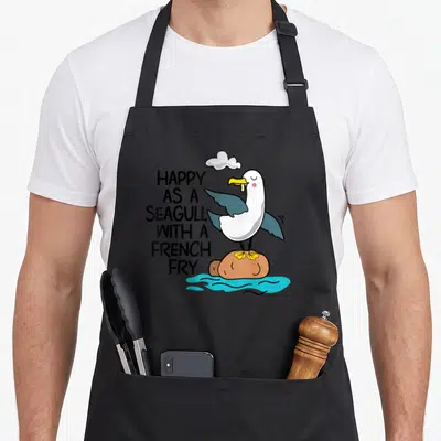 Seagull french apron - Cartoon seagull holding french fry standing on potato rock next to stacked black text happy as — shown on a black cotton canvas apron against a white background