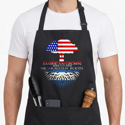 Caribbean american flag apron - Tree shaped like US flag above text American Grown With Nicaraguan Roots over roots — shown on a black cotton canvas apron against a white background
