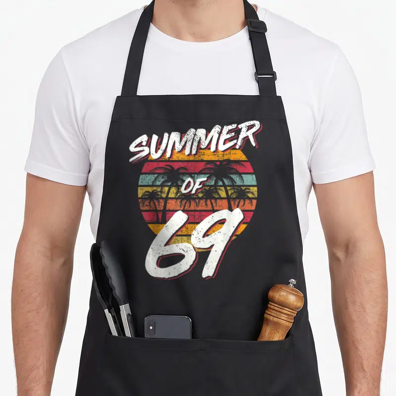 55th daddy apron - Retro Summer of 69 striped sunset with palm tree silhouettes and distressed white lettering — shown on a black cotton canvas apron against a white background