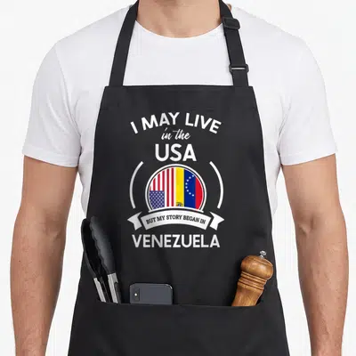 Began story of the apron - Stacked text I MAY LIVE in the USA BUT MY STORY BEGAN IN VENEZUELA around two flags — shown on a black cotton canvas apron against a white background