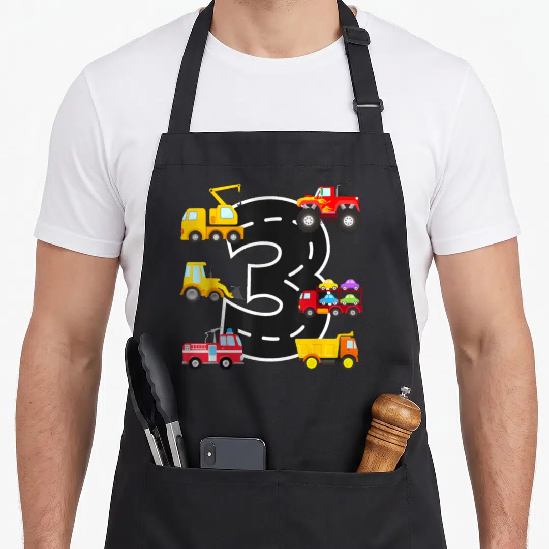 3rd birthday truck apron - Black number 3 road with fire truck, monster truck, and digger in cartoon style — shown on a black cotton canvas apron against a white background