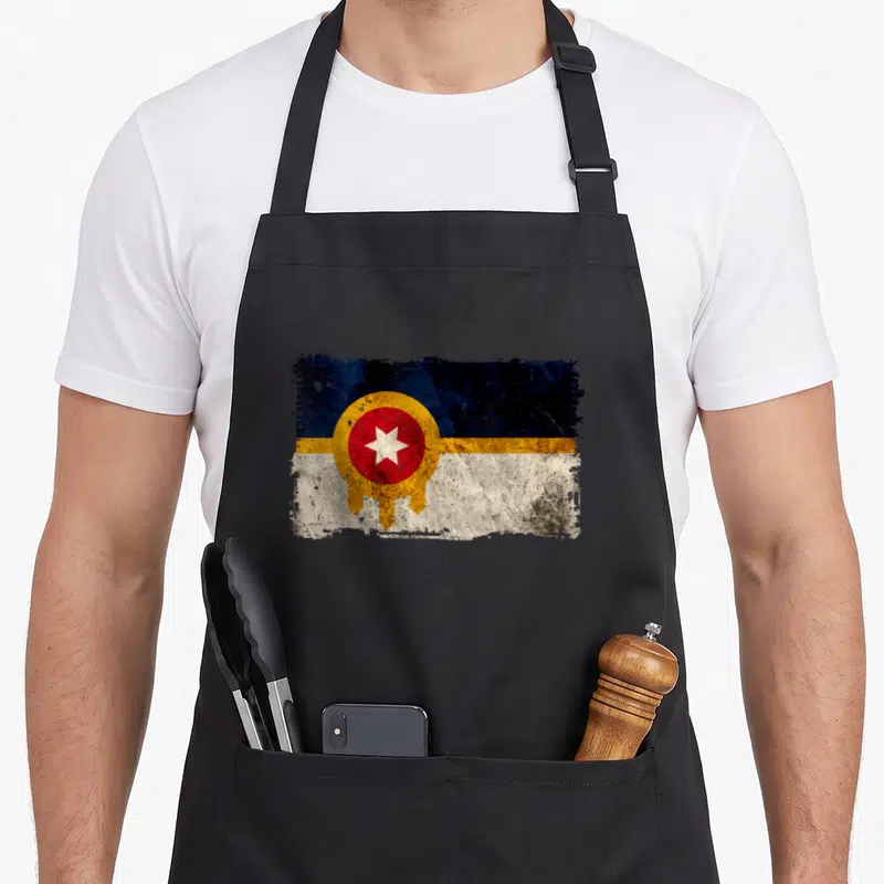 Tulsa distressed apron - Weathered Oklahoma state flag with blue yellow white red distressed texture on black fabric — shown on a black cotton canvas apron against a white background