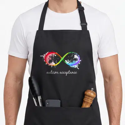 Cute instead autism acceptance apron - Rainbow infinity symbol with red yellow green blue white splatters and autism — shown on a black cotton canvas apron against a white background