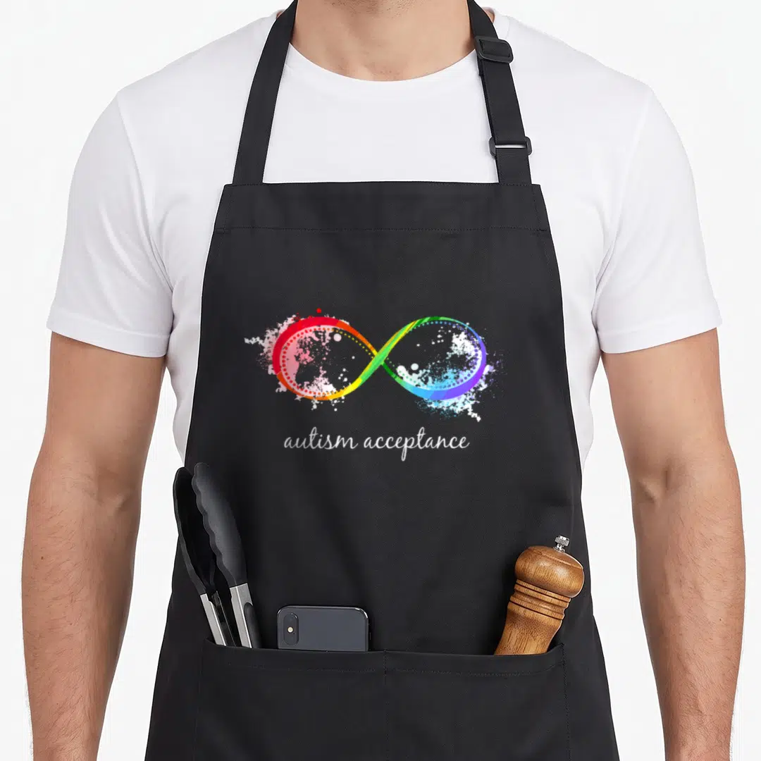 Cute instead autism acceptance apron - Rainbow infinity symbol with red yellow green blue white splatters and autism — shown on a black cotton canvas apron against a white background