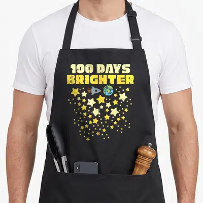 Brighter star apron - 100 DAYS BRIGHTER text above rocket earth and scattered yellow grey stars — shown on a black cotton canvas apron against a white background