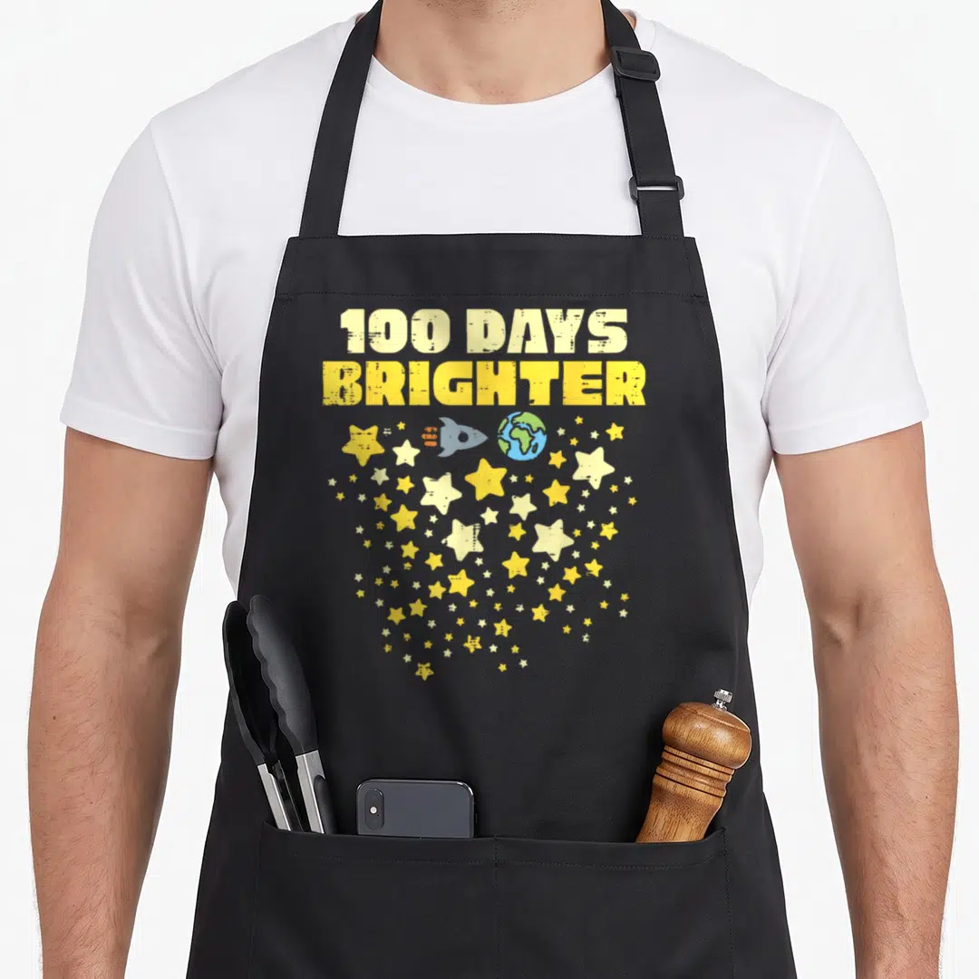 Brighter star apron - 100 DAYS BRIGHTER text above rocket earth and scattered yellow grey stars — shown on a black cotton canvas apron against a white background