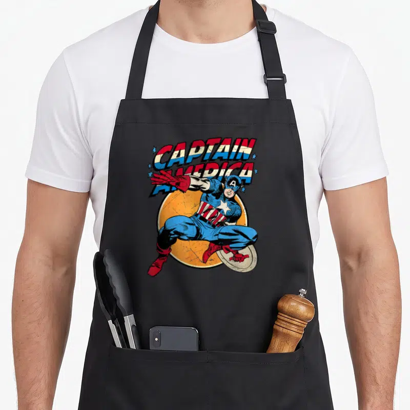 Cute comic hobby apron - Diving Captain America holding shield in front of orange circle with distressed red white blue — shown on a black cotton canvas apron against a white background