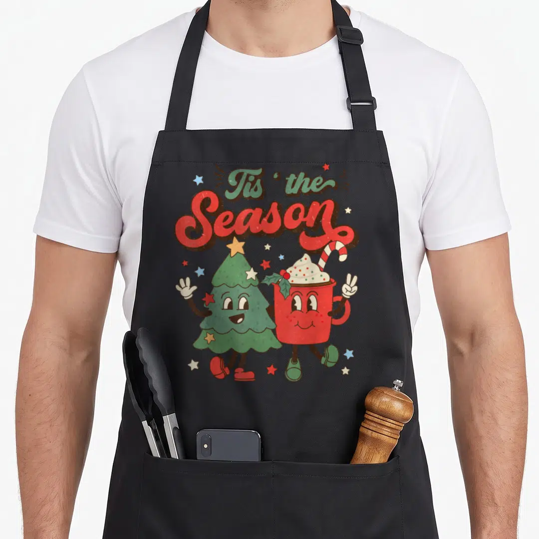 Tis christmas tree apron - Cartoon Christmas tree and red cocoa mug holding hands under Tis the Season text — shown on a black cotton canvas apron against a white background