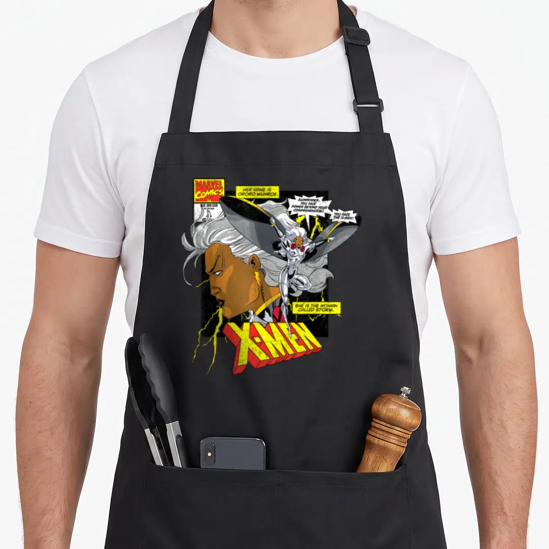 Called hobby apron - X-Men Storm comic cover with profile portrait, floating figure, and bold yellow red X-MEN lettering — shown on a black cotton canvas apron against a white background