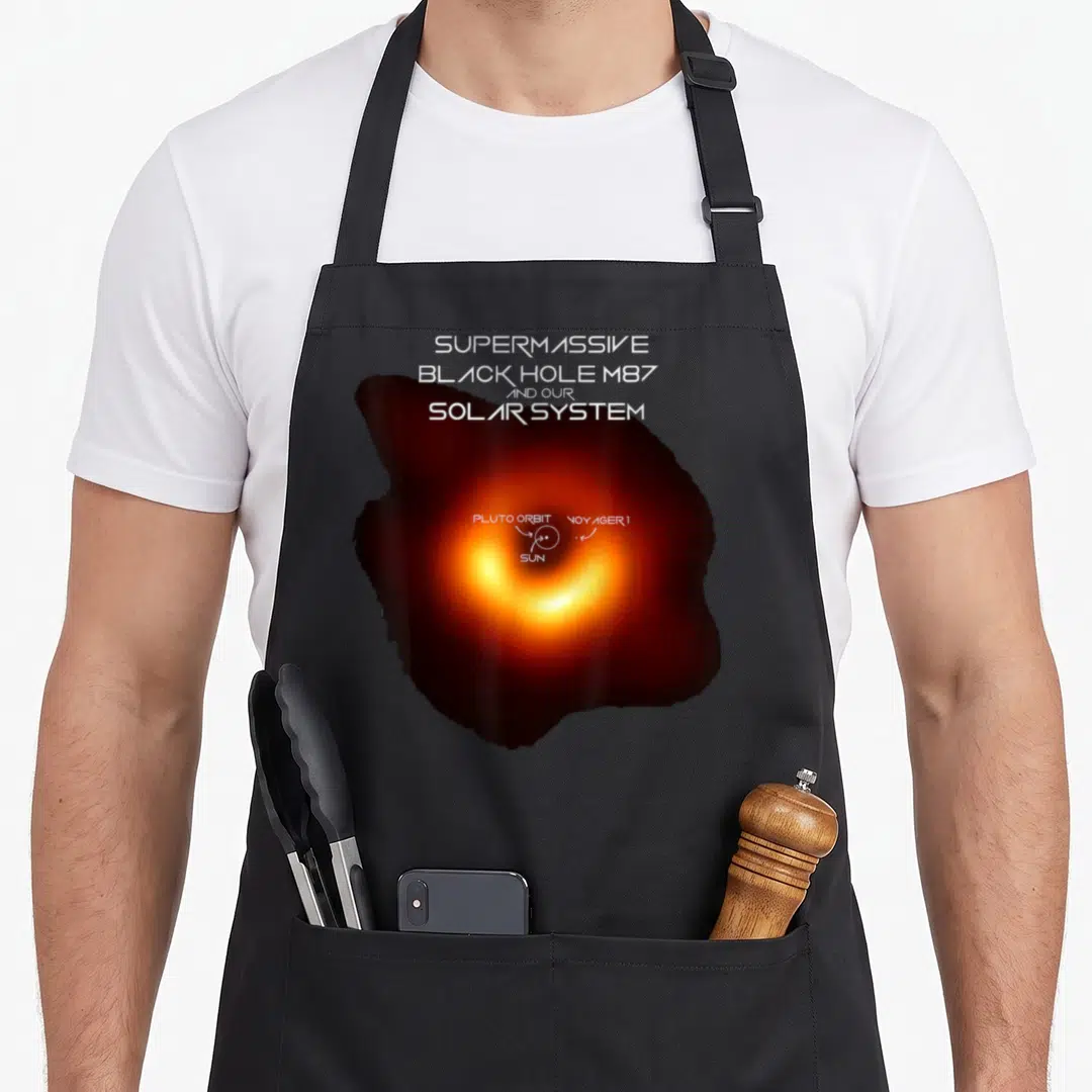 M87 hole apron - Realistic orange black hole with overlaid solar system diagram labels Sun and Voyager 1 — shown on a black cotton canvas apron against a white background