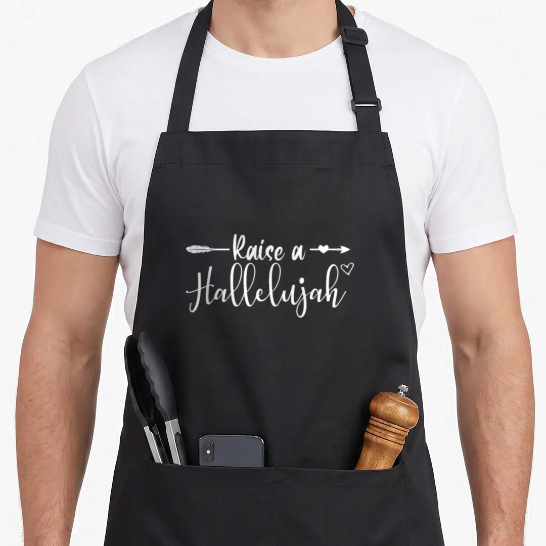 Raise congratulations apron - White Raised a Hallelujah text with feathered arrow and heart icons on black fabric — shown on a black cotton canvas apron against a white background