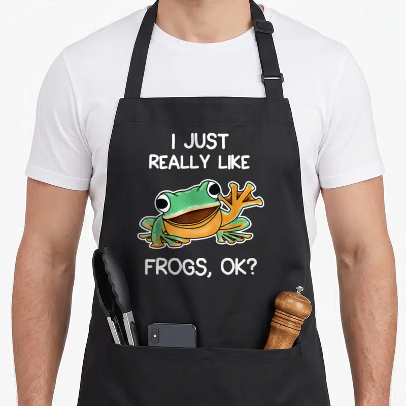 Owner frog aprons - Cartoon green frog with orange belly between white text reading I Just Really Like Frogs Ok — shown on a black cotton canvas apron against a white background