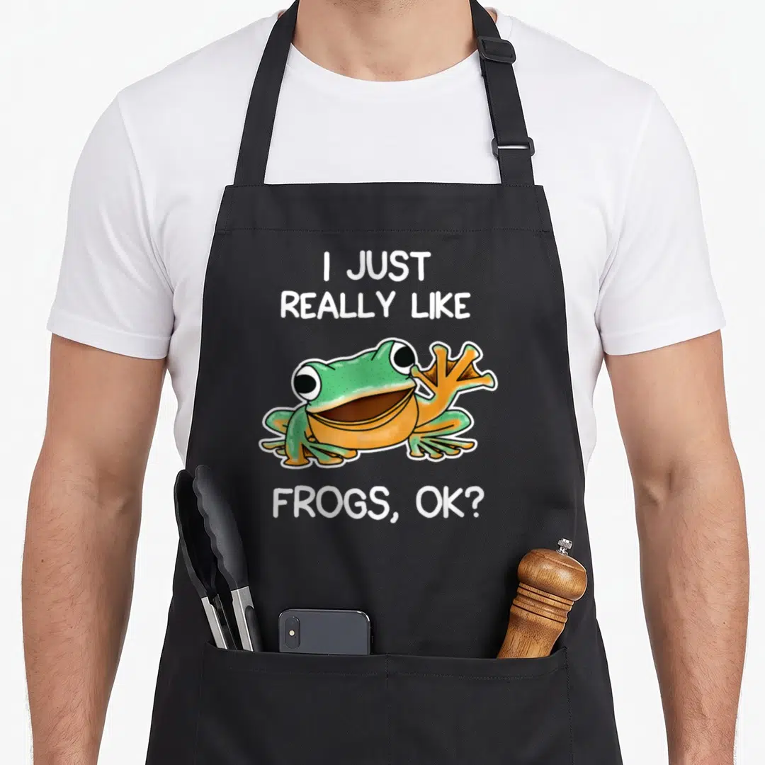 Owner frog aprons - Cartoon green frog with orange belly between white text reading I Just Really Like Frogs Ok — shown on a black cotton canvas apron against a white background