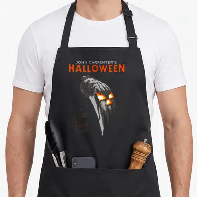 Came horror movie apron - Halloween knife overlapping pumpkin with orange and grey lettering on dark background — shown on a black cotton canvas apron against a white background