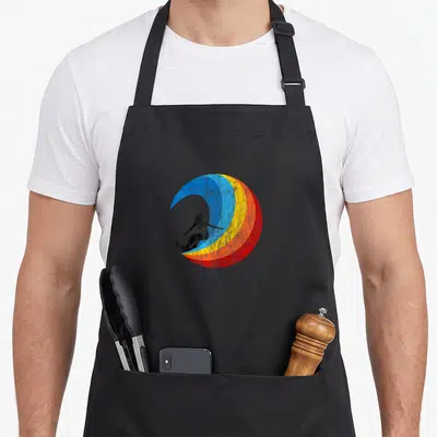 Surfer summer mode apron - Black surfer silhouette on blue yellow orange and red crescent wave — shown on a black cotton canvas apron against a white background