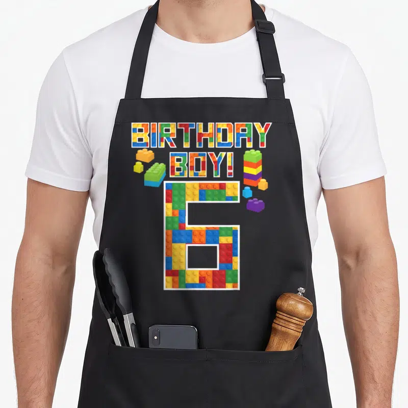 6th building aprons - BIRTHDAY BOY! 6 text filled with colorful plastic brick patterns and loose blocks — shown on a black cotton canvas apron against a white background