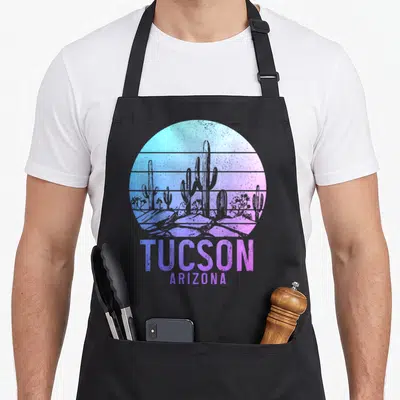 Tucson cactus apron - Circular desert landscape with cacti rocks and stacked text TUCSON ARIZONA in blue purple pink — shown on a black cotton canvas apron against a white background