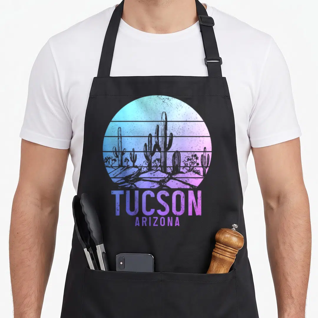 Tucson cactus apron - Circular desert landscape with cacti rocks and stacked text TUCSON ARIZONA in blue purple pink — shown on a black cotton canvas apron against a white background
