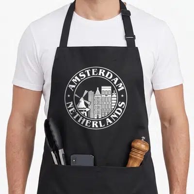 Holland netherlands apron - White circular emblem with Amsterdam skyline, windmill, buildings, and two stars — shown on a black cotton canvas apron against a white background