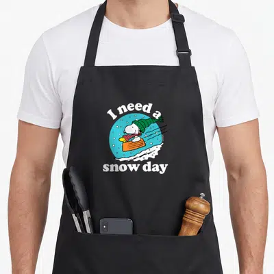 Snow christmas apron - Snoopy in green hat on orange sled in blue circle with white text — shown on a black cotton canvas apron against a white background
