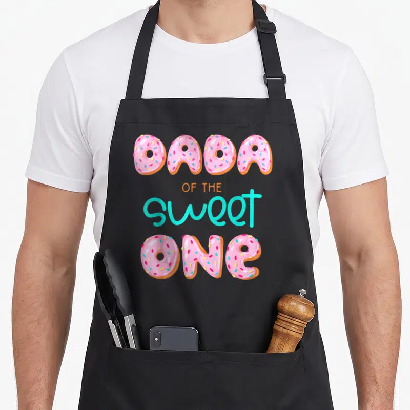 Dada family aprons - Pink donuts spelling DADA and ONE with orange and turquoise text — shown on a black cotton canvas apron against a white background