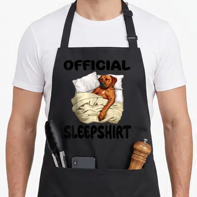 Ridgeback dog apron - Cartoon dog lying in bed with pillows and blanket labeled OFFICIAL SLEEPSHIRT — shown on a black cotton canvas apron against a white background