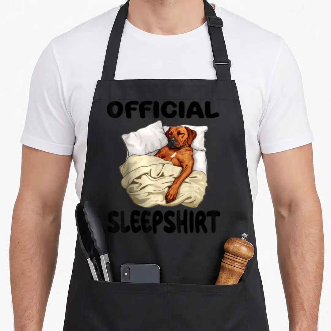 Ridgeback dog apron - Cartoon dog lying in bed with pillows and blanket labeled OFFICIAL SLEEPSHIRT — shown on a black cotton canvas apron against a white background