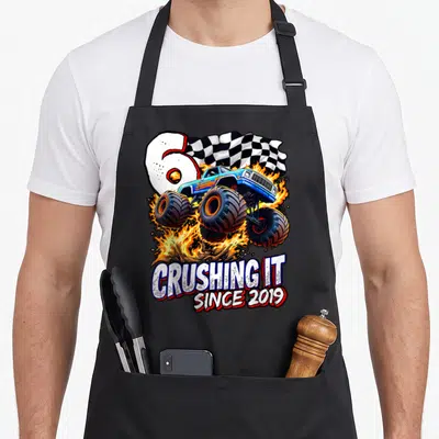6th truck apron - Blue monster truck jumping through fire with number 6 and CRUSHING IT SINCE 2019 text — shown on a black cotton canvas apron against a white background