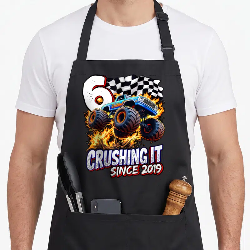 6th truck apron - Blue monster truck jumping through fire with number 6 and CRUSHING IT SINCE 2019 text — shown on a black cotton canvas apron against a white background