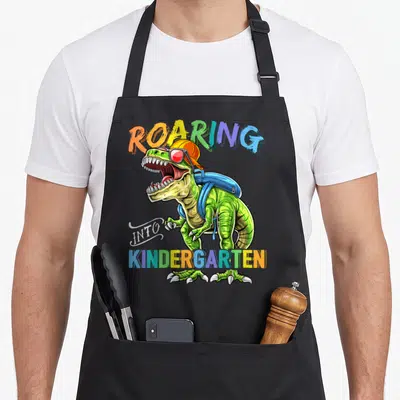 Roaring kindergarten dinosaur apron - T Rex wearing orange cap and blue backpack with ROARING INTO KINDERGARTEN text — shown on a black cotton canvas apron against a white background