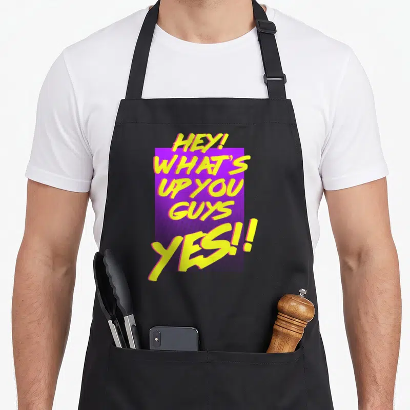 Dawson hey novelty apron - Yellow brush script text HEY WHATS UP YOU GUYS YES over purple rectangle — shown on a black cotton canvas apron against a white background