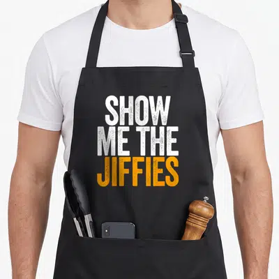 Show swagazon apron - Stacked white and orange distressed text reading SHOW ME THE JIFFIES on black fabric — shown on a black cotton canvas apron against a white background