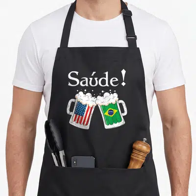 Saude toast apron - Two clinking beer mugs with US and Brazil flags under white Saúde text — shown on a black cotton canvas apron against a white background