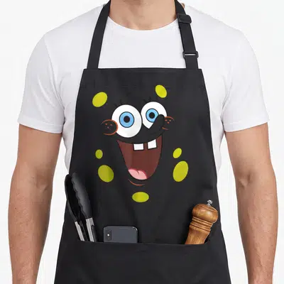 Face halloween white apron - Cartoon face with blue eyes and buck teeth surrounded by yellow ovals on grey — shown on a black cotton canvas apron against a white background
