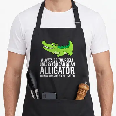 Crocodile unless apron - Green cartoon alligator above white text ALWAYS BE YOURSELF UNLESS YOU CAN BE AN ALLIGATOR — shown on a black cotton canvas apron against a white background